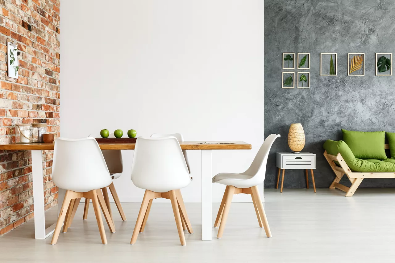 Contemporary interior design of dining space with communal table against brick wall and open living room with simple sofa and plant decorations on textured gray wall