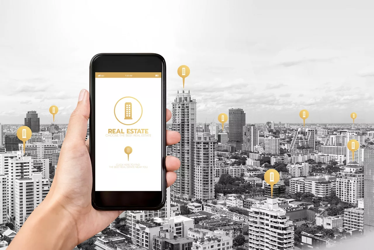 Hand holding smartphone with application to find  real estate on screen and buildings in the city as background