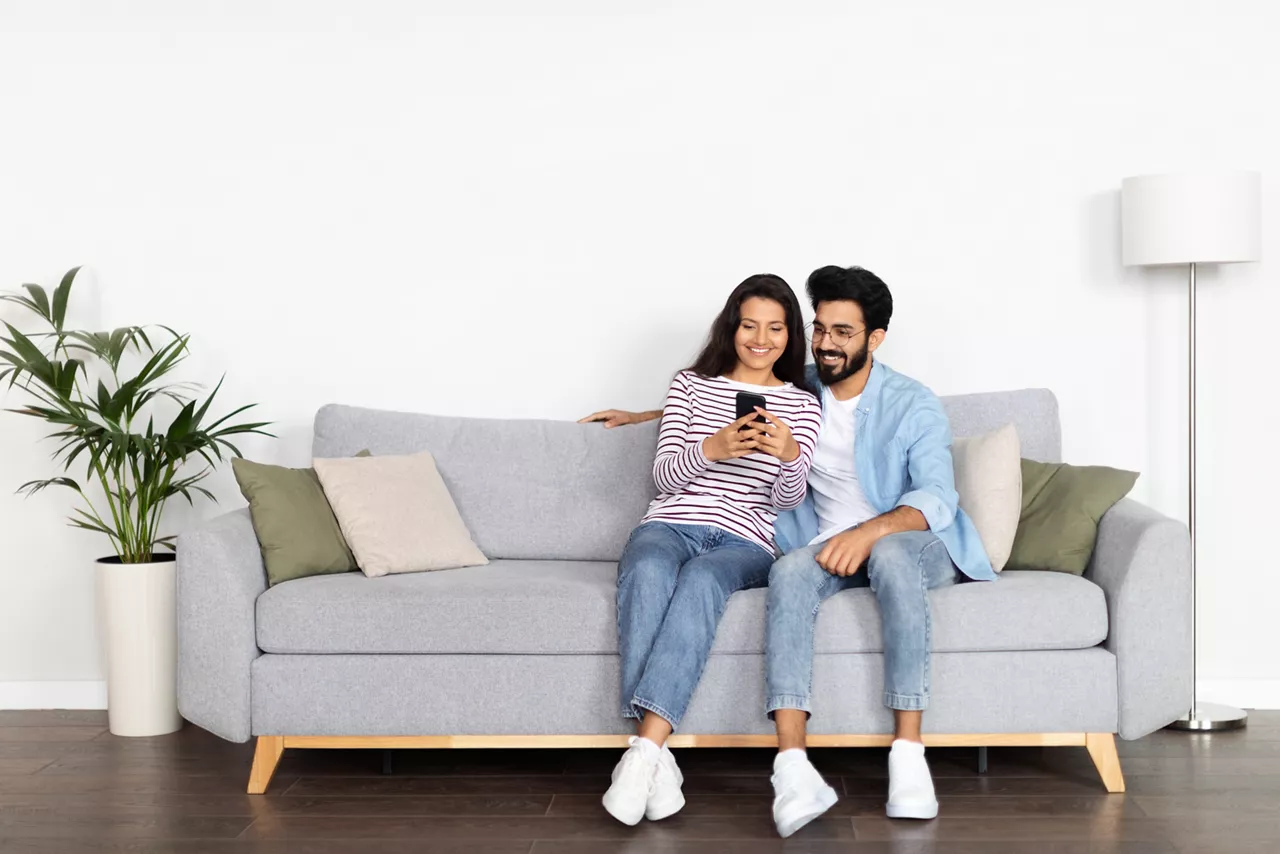 Relaxed cheerful beautiful young multicultural couple middle eastern guy and indian woman sitting on sofa at home, using smartphone and smiling, shopping online, surfing on Internet, copy space