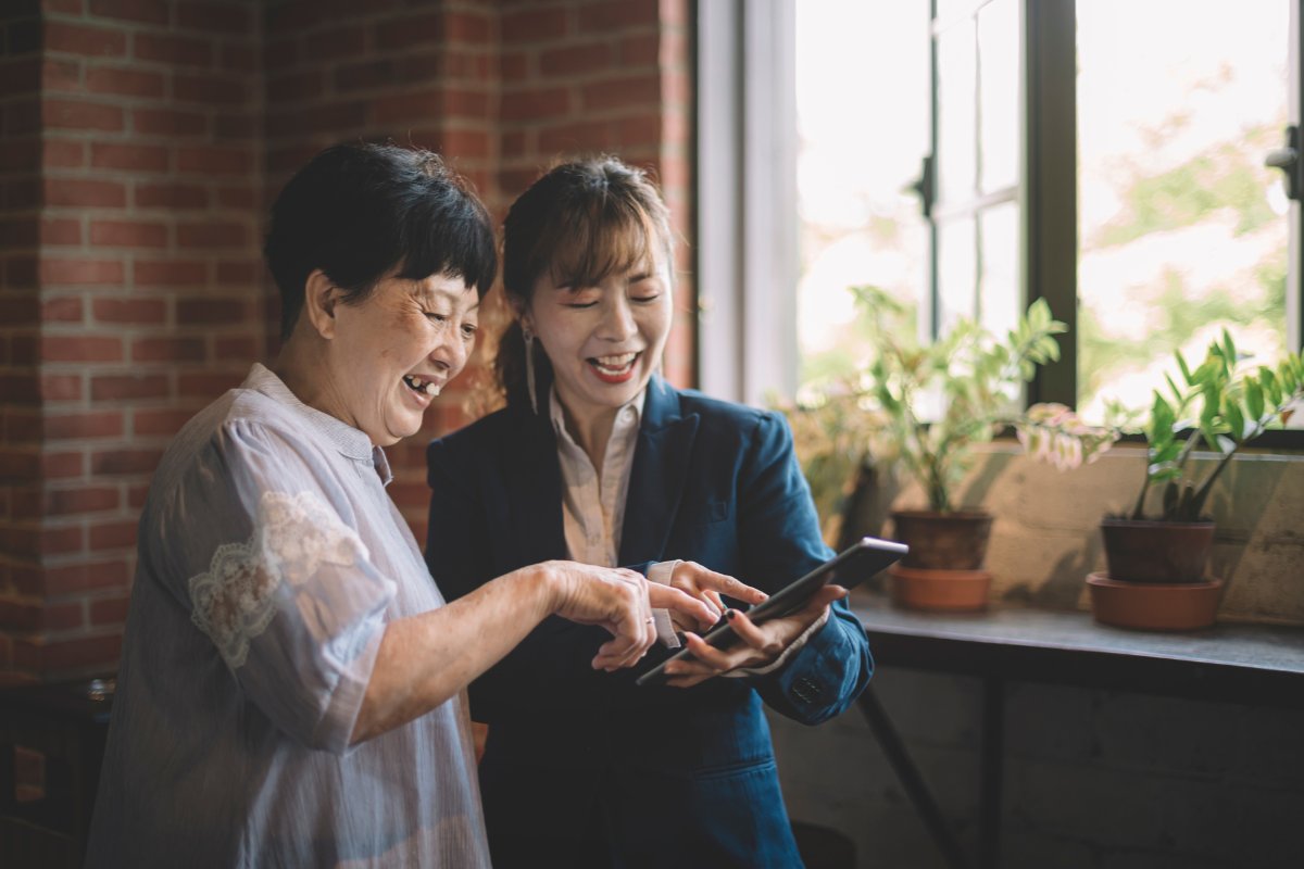 An old mother holds an tablet computer mini sitting on sofa. She and her daughter watch some photos and smile together.