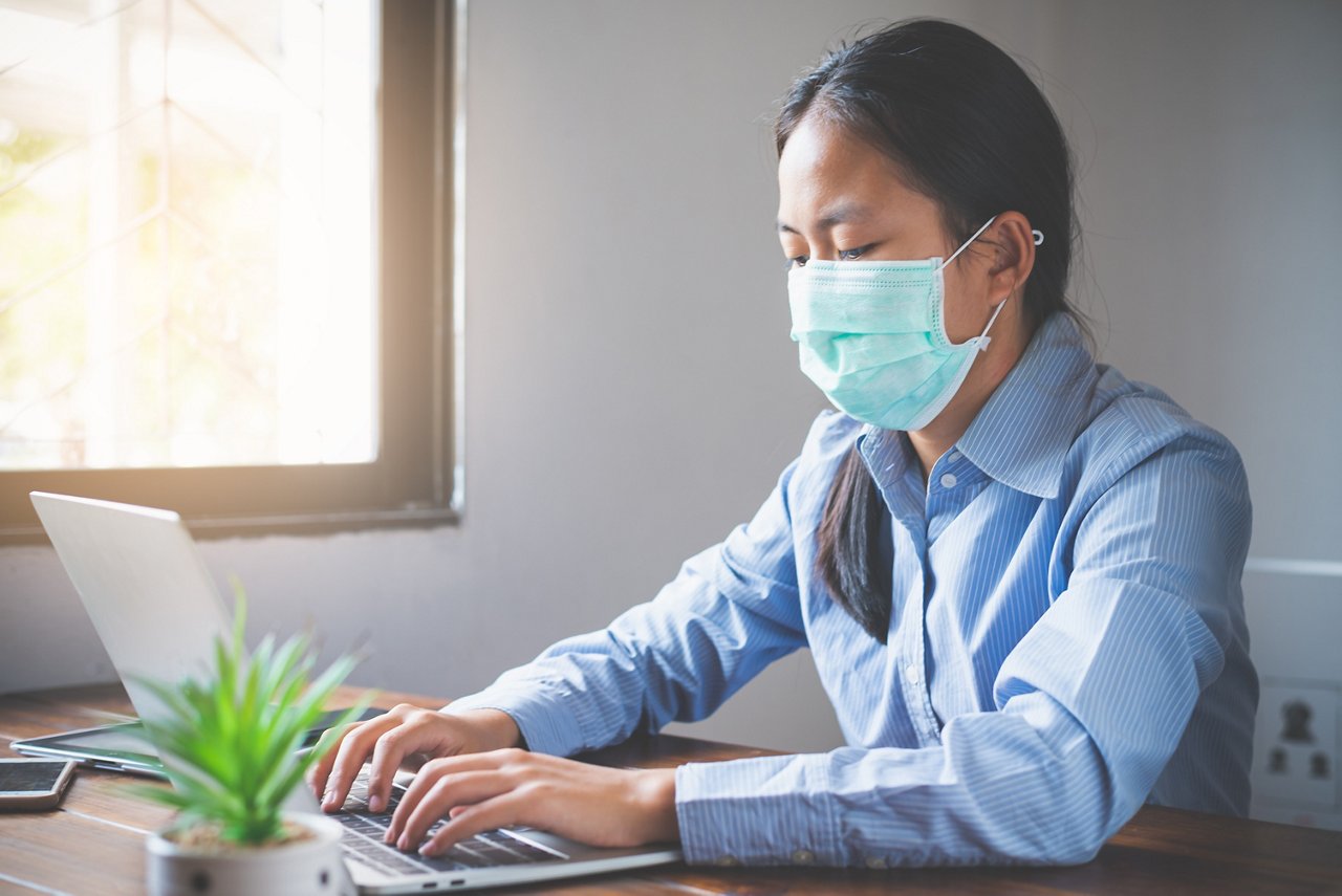 A woman in office attire and a face mask types on her lapto