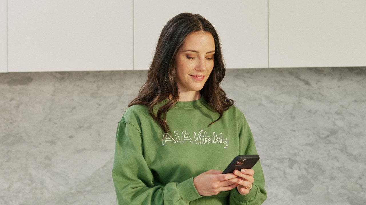 A woman in a green sweatshirt gazes at her phone, appearing focused and engaged with the screen.