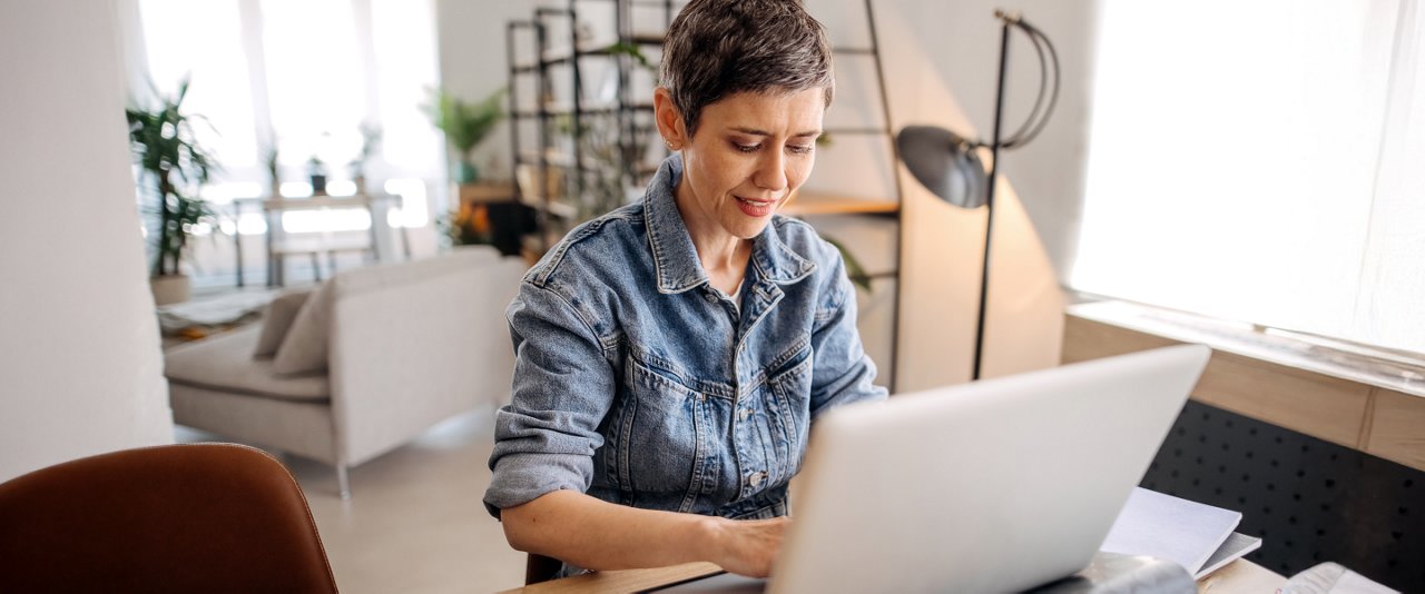 A woman sits at a desk, focused on her laptop, with a notepad and pen beside her.