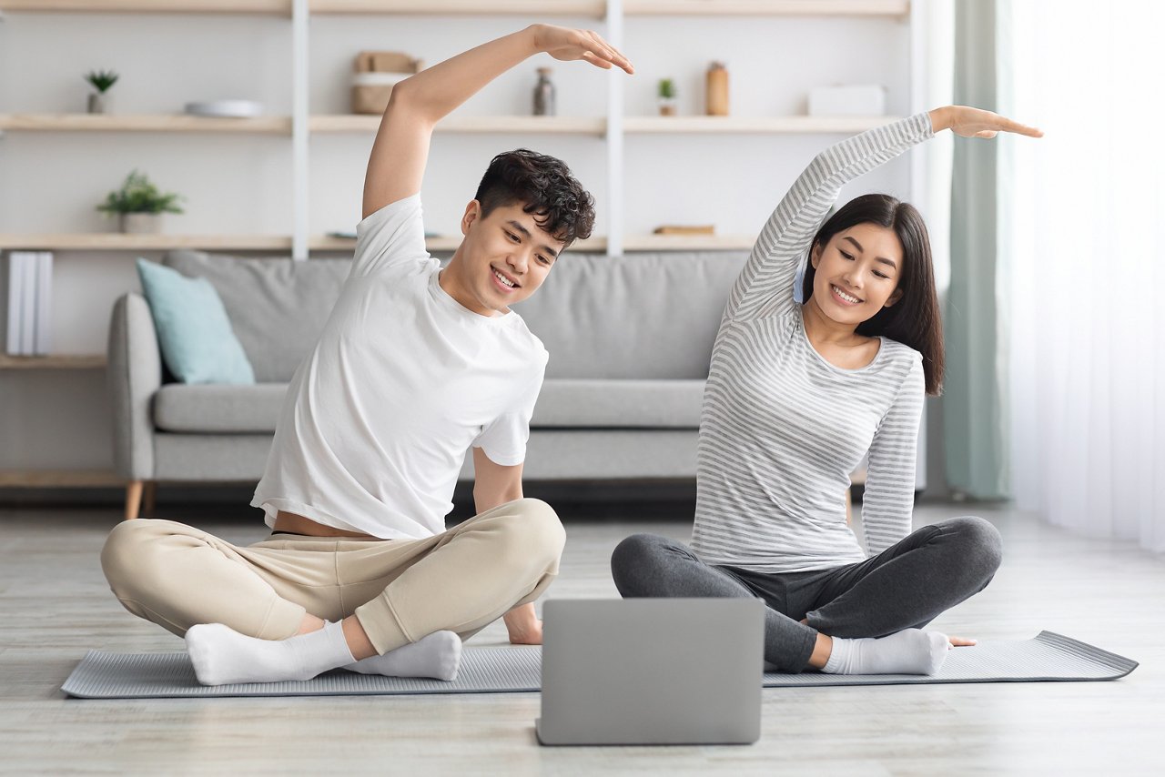 Cheerful asian family practicing yoga at home, stretching on fitness mat, looking at laptop screen, having online class, living room interior, copy space. Sporty black couple exercising together
