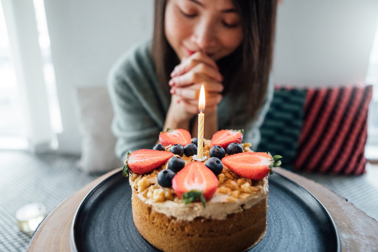 It's party and celebration time! Young Asian woman is making a wish in front of a beautiful coffee & walnut flavoured Victoria sponge birthday cake in living room with cushions on the floor.