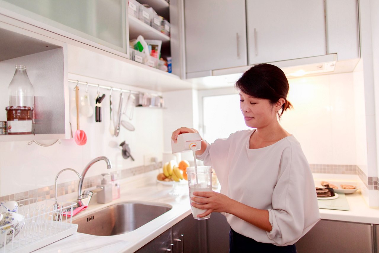 An Asian woman pours milk in a glass in her kitchen.