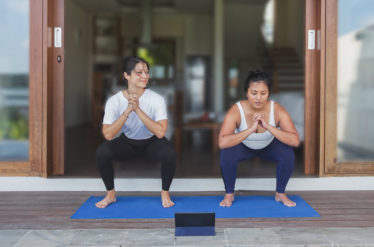 Solo Asian man performs a squat on a mat near the gym's windows. 