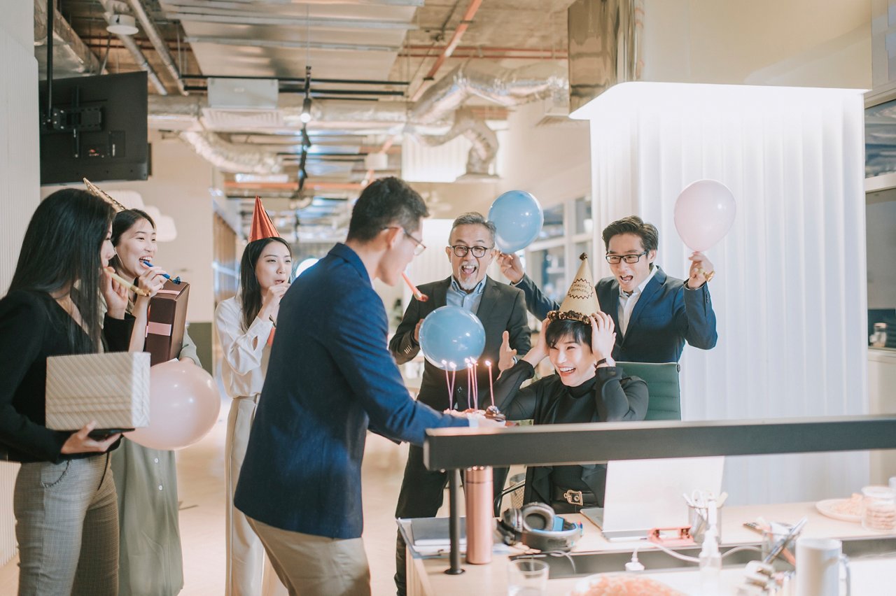 Office workers surprise a colleague at their workplace with birthday cake and balloons.