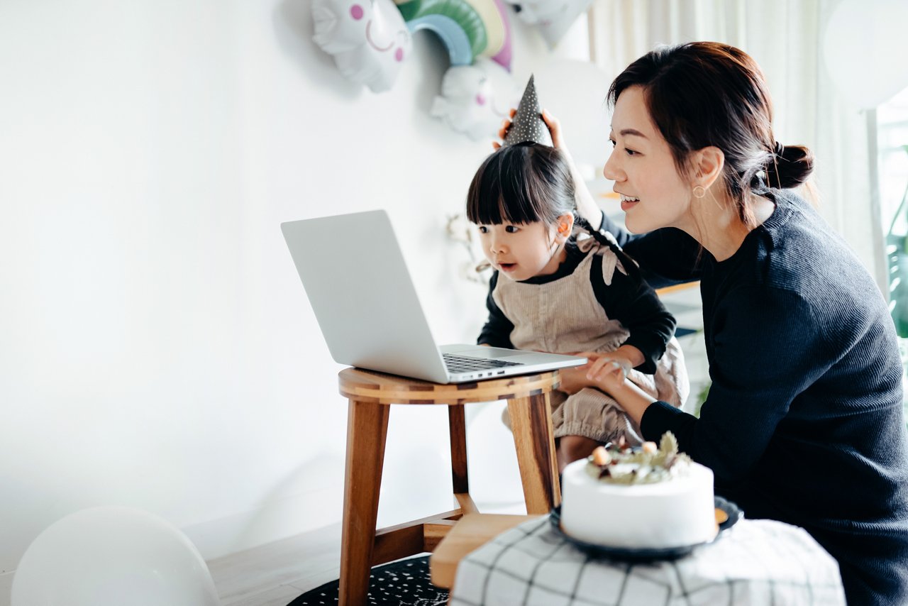 Lovely Asian girl and mother celebrating birthday with friends / family by having a virtual birthday party at home. Enjoying birthday celebration in front of the laptop during pandemic. Practicing social distancing. Birthday lifestyle theme