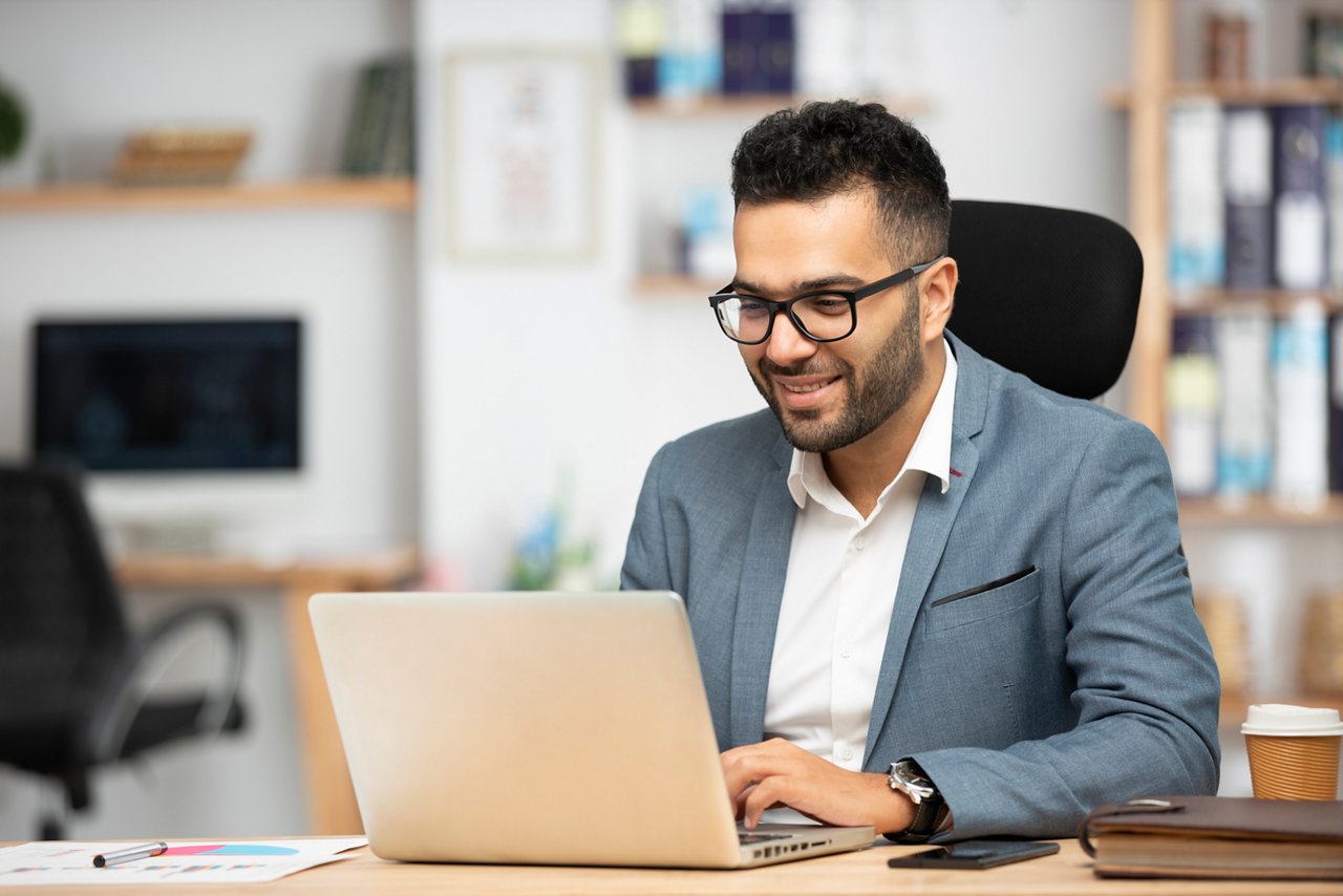 A bearded man wearing a suit is seen typing on his laptop at an office