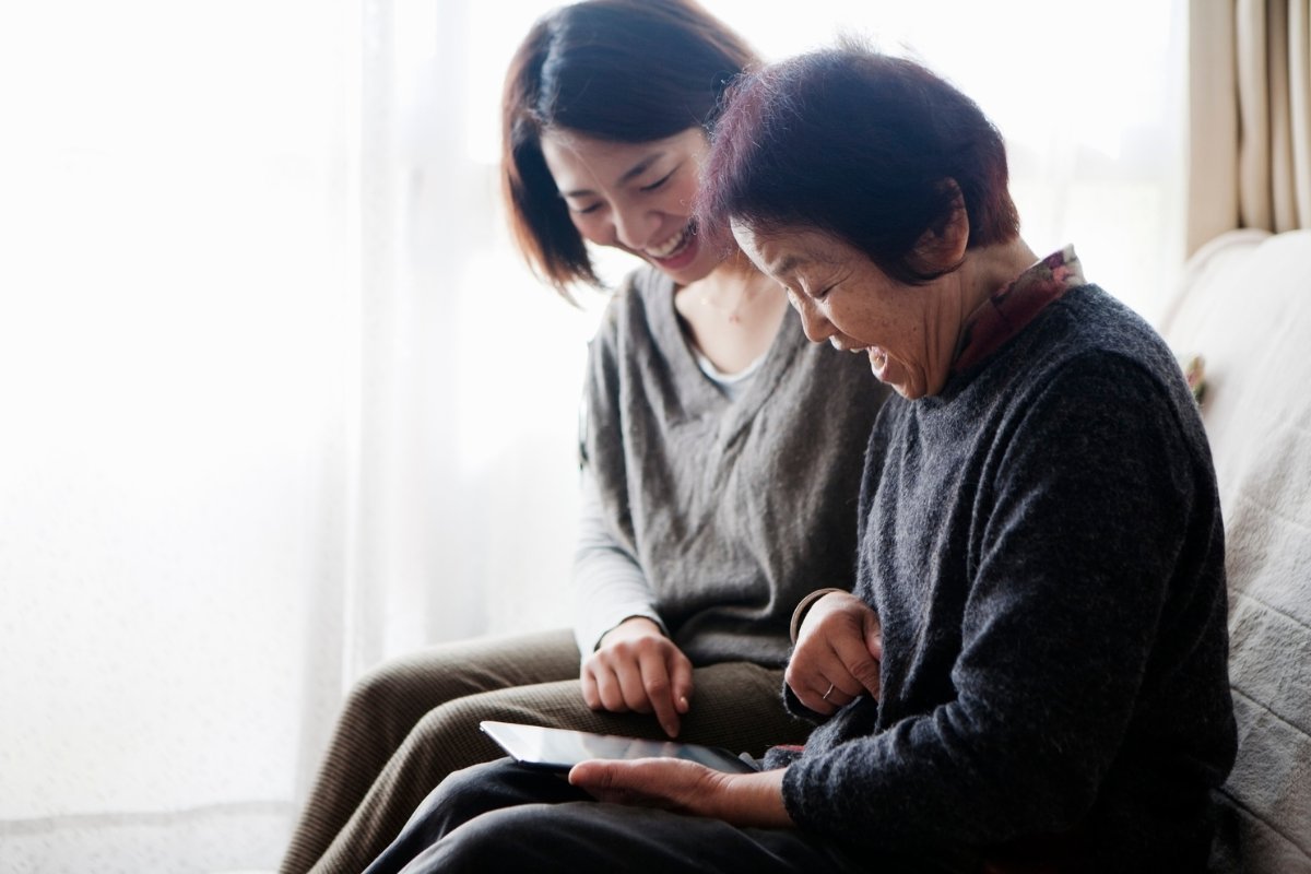 An old mother holds an tablet computer mini sitting on sofa. She and her daughter watch some photos and smile together.