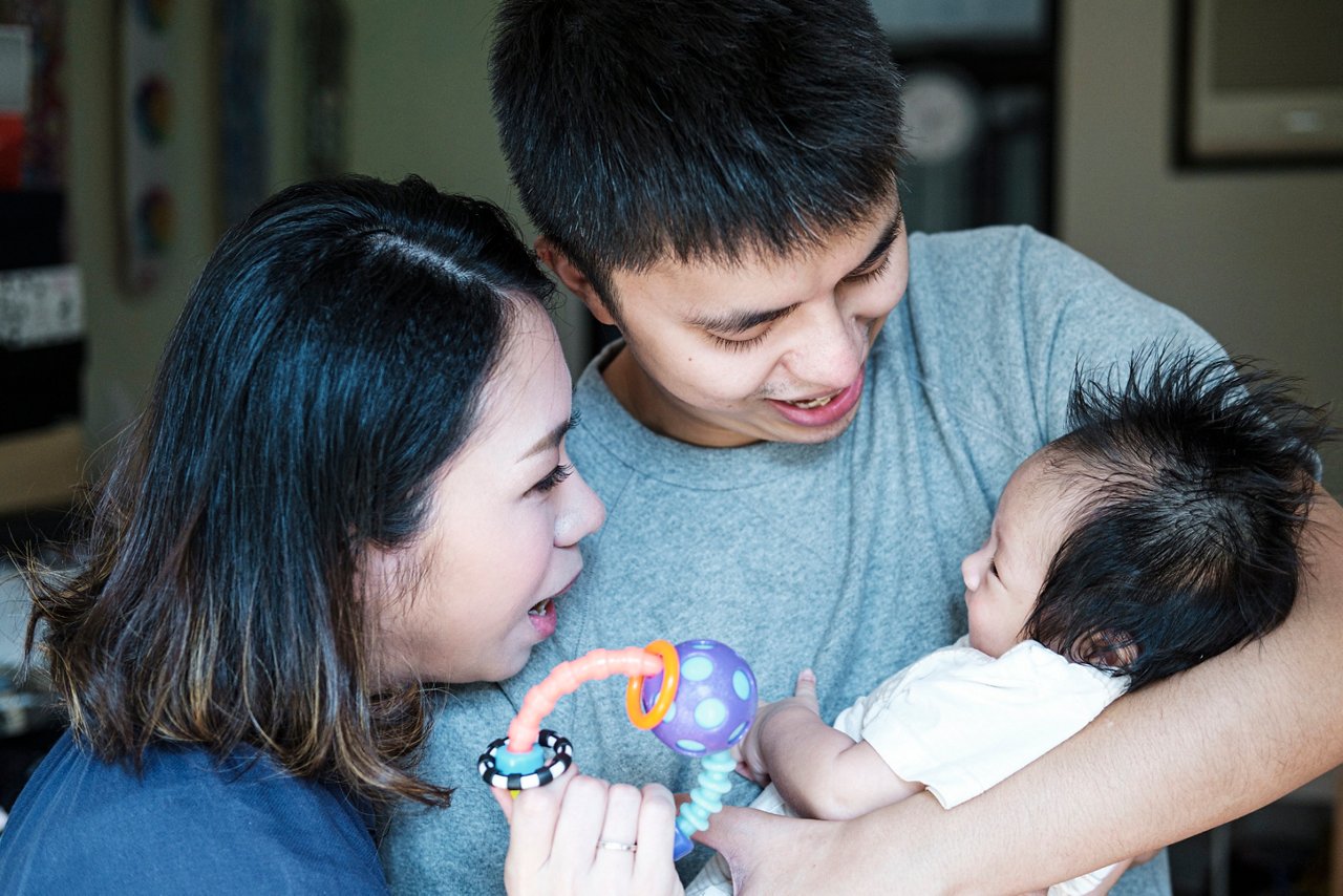 A father is holding his baby while the mother is holding a baby rattle toy