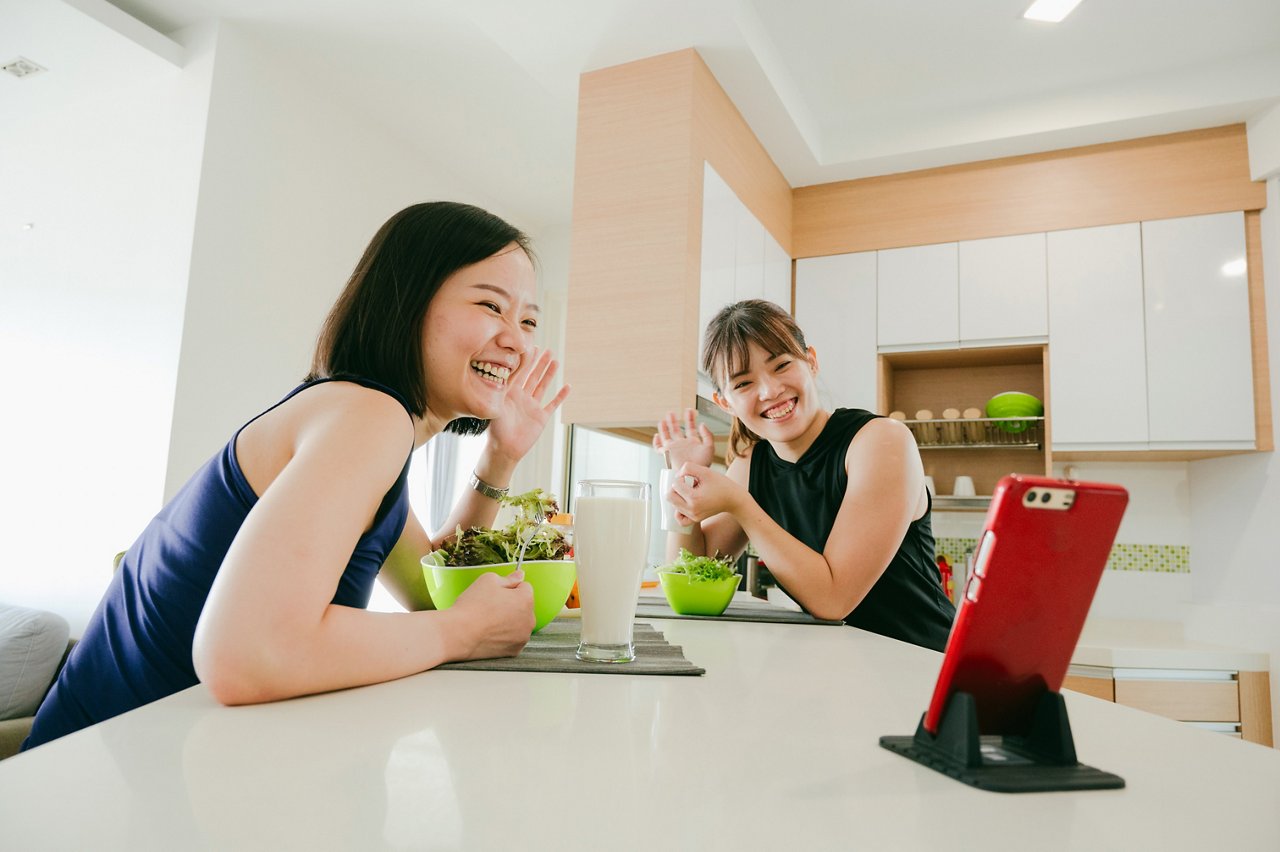 Two young Asian women eat salad in the kitchen and talk to a friend on a mobile phone