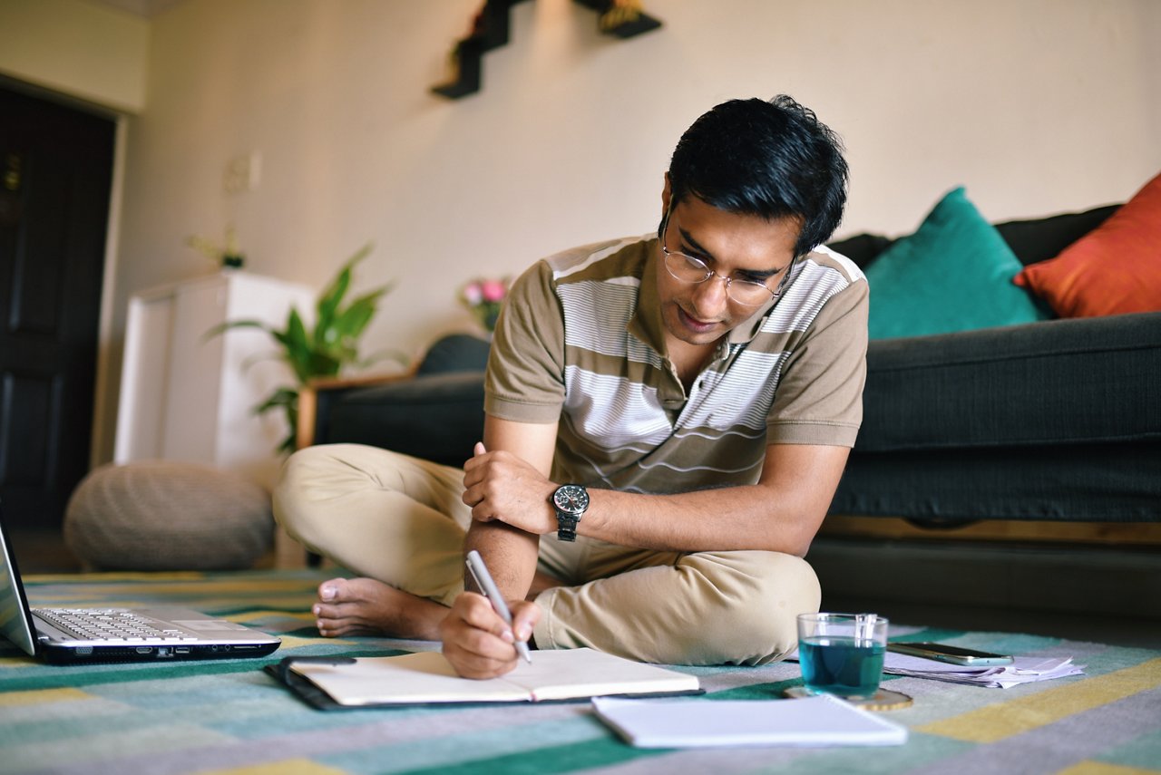 A man with glasses writing on a notebook with his laptop beside him