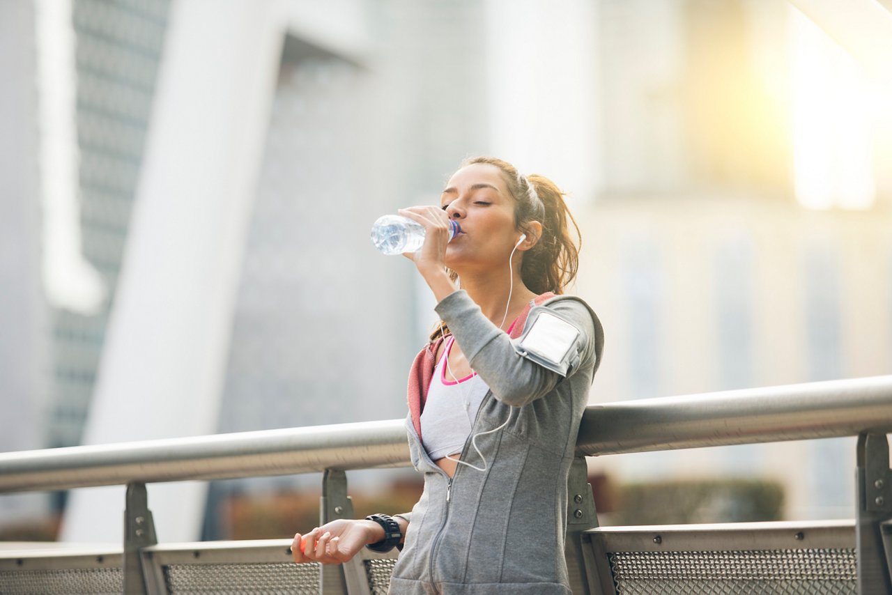 Woman in workout gear is drinking water from a bottle as she listens to music with her earphones
