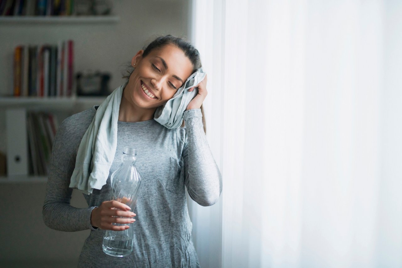 A woman wiping her sweat with a towel and holding a water bottle after working out