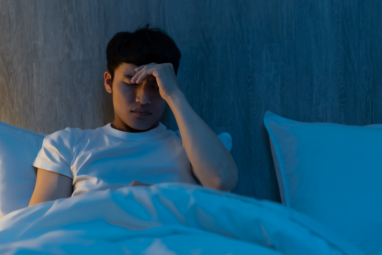 Young man sitting on a bed holding his forehead, looking tired and unwell.