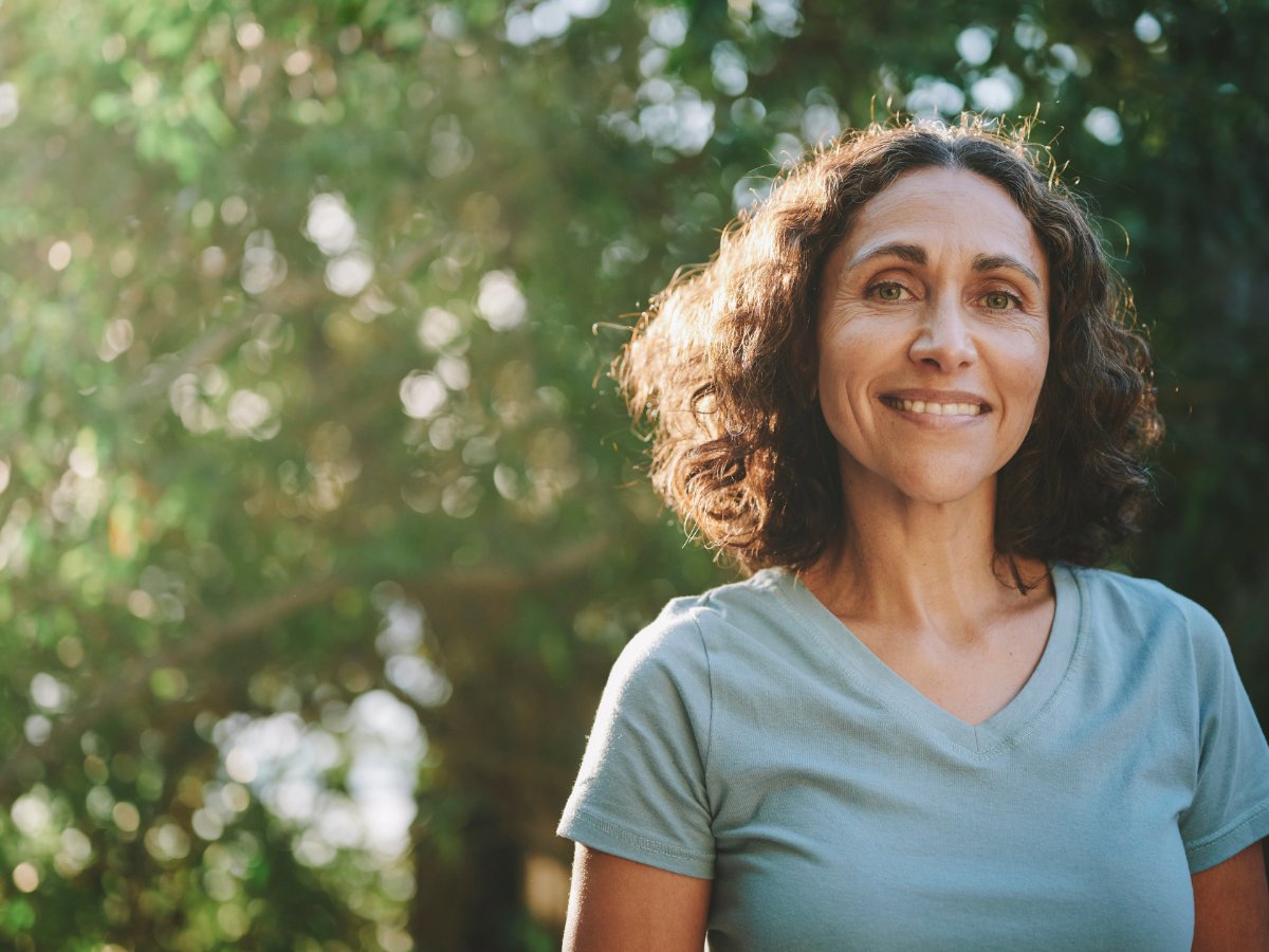 A woman smiles joyfully while standing amidst a backdrop of lush green trees.