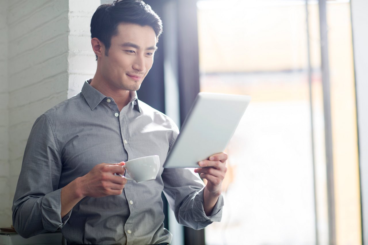 一位精明的男士拿著一杯咖啡並在使用平板電腦，查看自己於友邦峻宇會可享用的財富管理服務禮遇。
