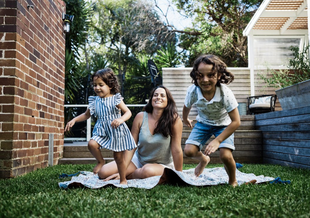 A woman and two children joyfully playing on a blanket in a sunny backyard, surrounded by grass and trees.