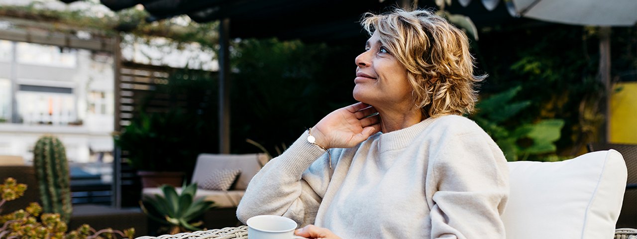 A woman relaxes on a patio, holding a cup of coffee, with a serene outdoor setting in the background