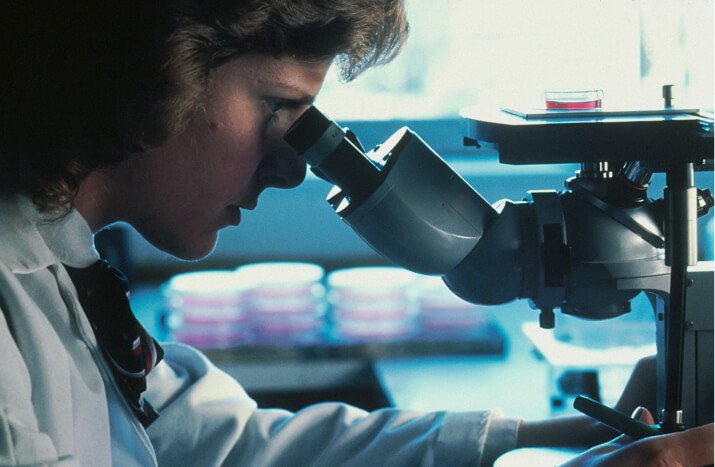 A woman in a lab coat intently examines a specimen through a microscope in a laboratory setting.