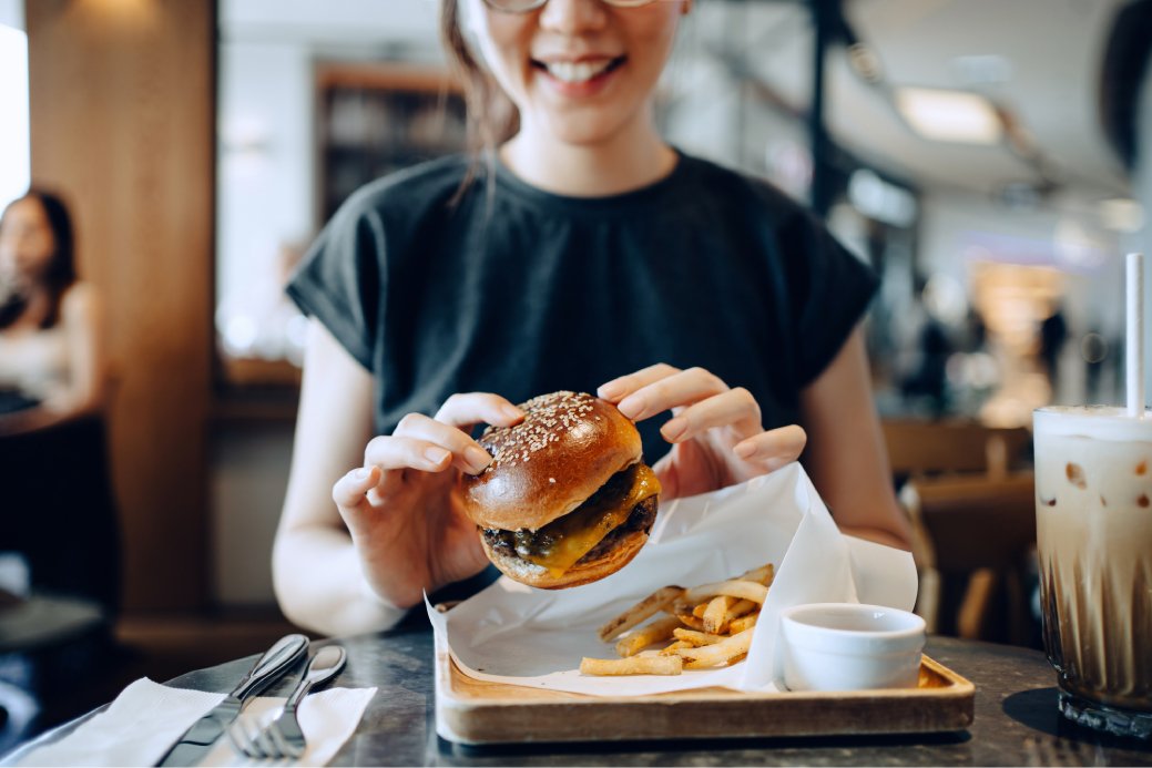 A smiling young Asian woman is enjoying freshly made delicious burger with fries and a glass of iced coffee in a café.