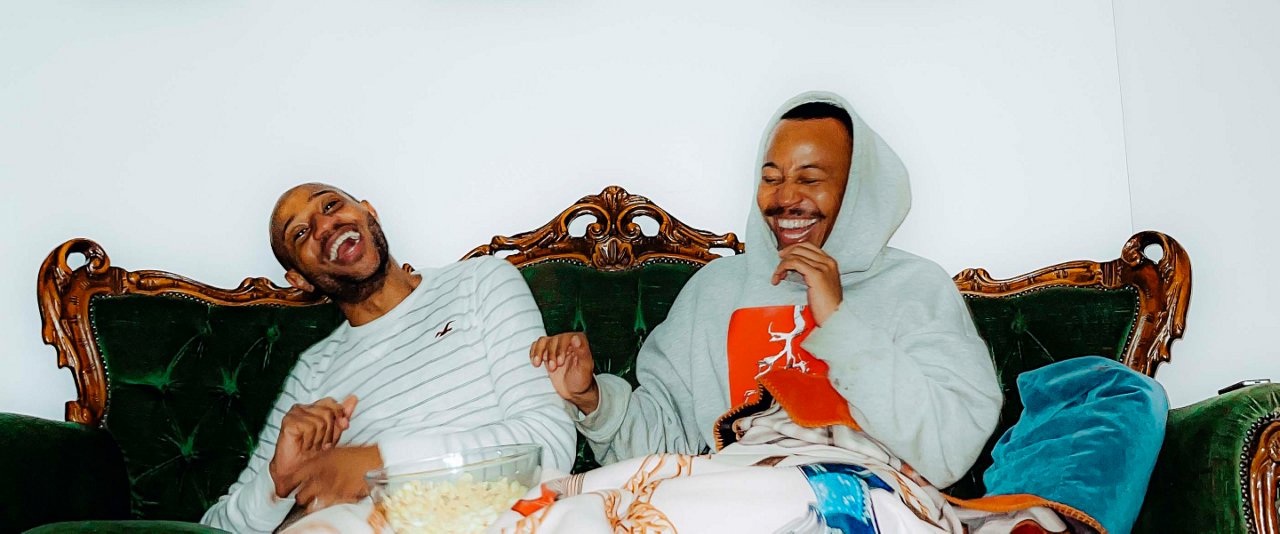 Two men sitting on a couch with a blanket draped over them, chatting and enjoying a cozy evening together.