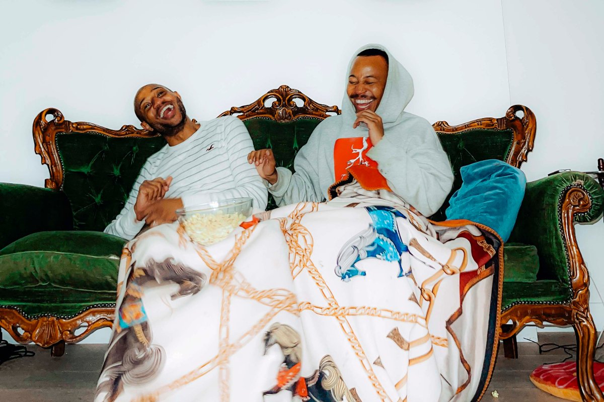 Two men sitting on a couch with a blanket draped over them, chatting and enjoying a cozy evening together.