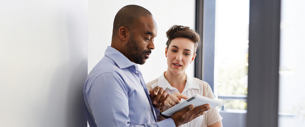 In the office, a woman and a man are talking while focusing on a tablet.