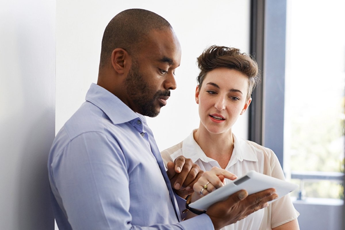 In the office, a woman and a man are talking while focusing on a tablet.