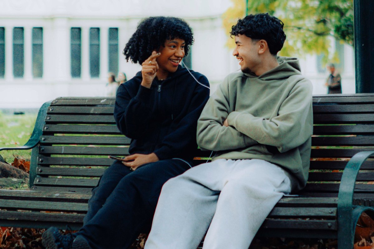 Friends seated on a park bench listening to each other's music with earbuds.