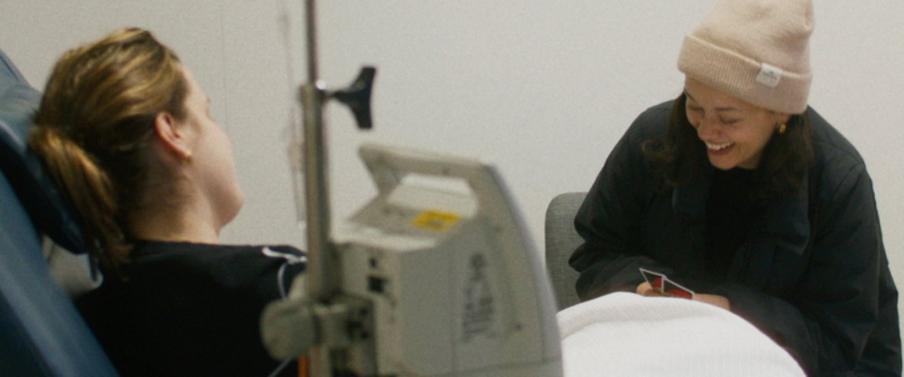 A woman lies in a hospital bed talking to a friend who is visiting.