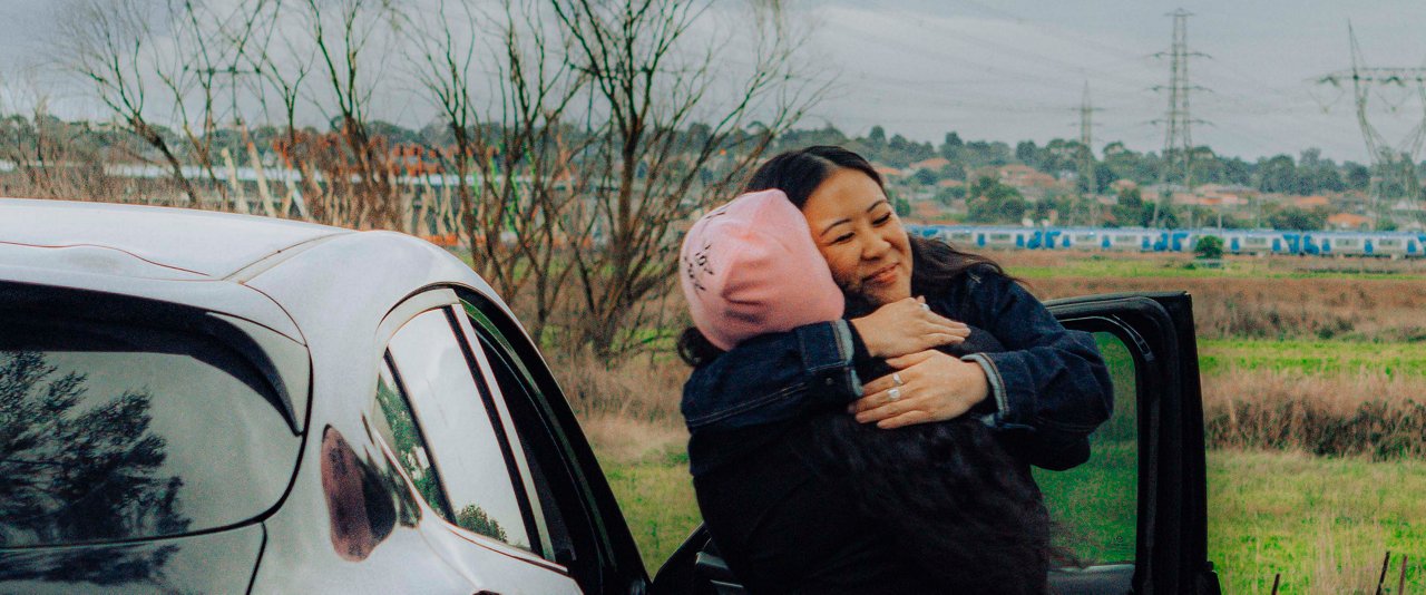 Two women hugging by a car, both looking happy.