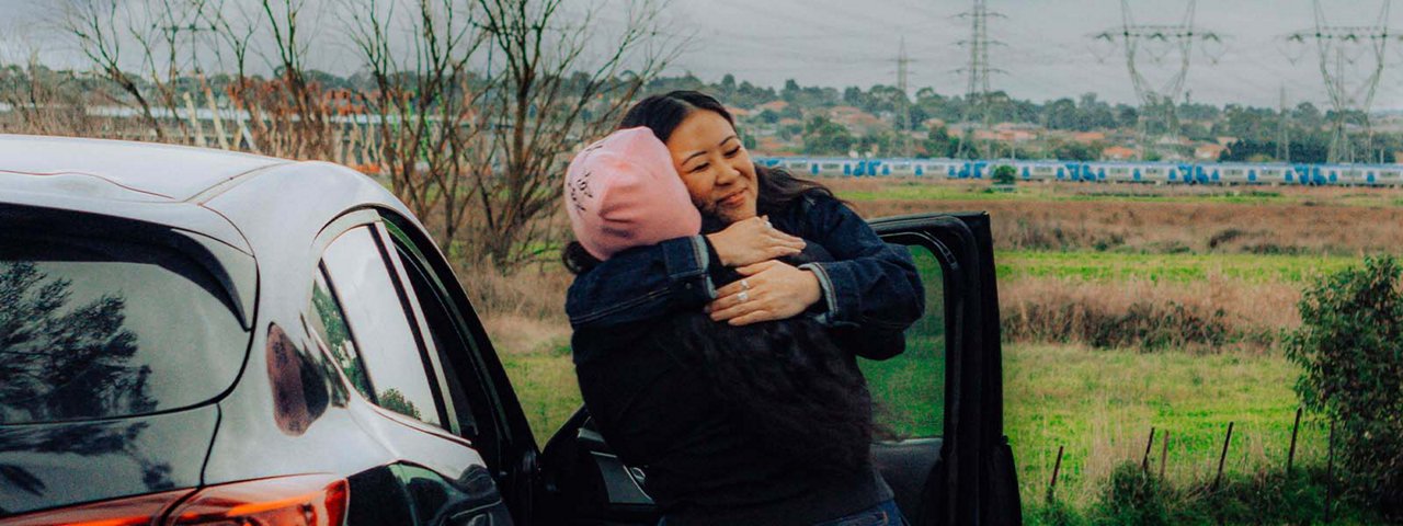 Two women hugging by a car, both looking happy.