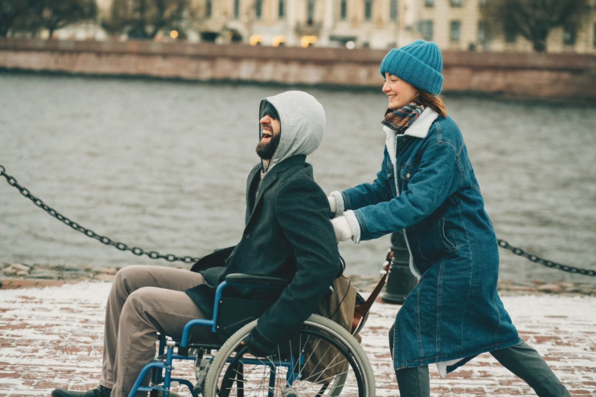 On a sidewalk, a woman pushes a man in a wheelchair.