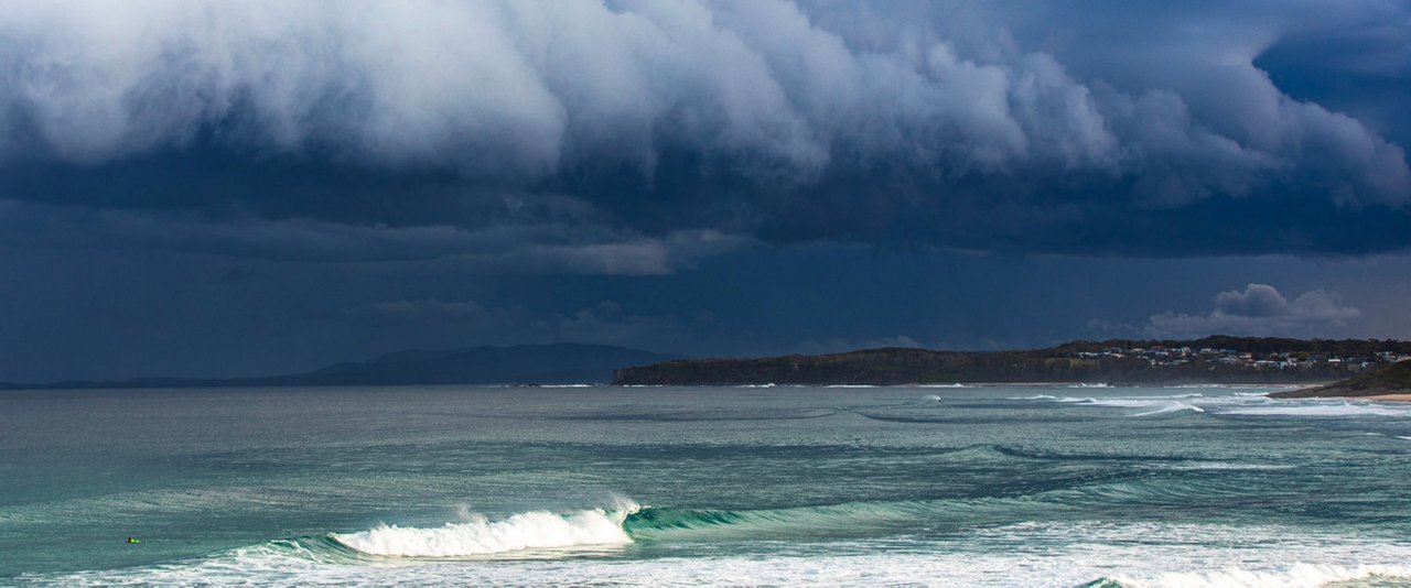 A person surfing on a board at the beach, with dark storm clouds looming overhead.