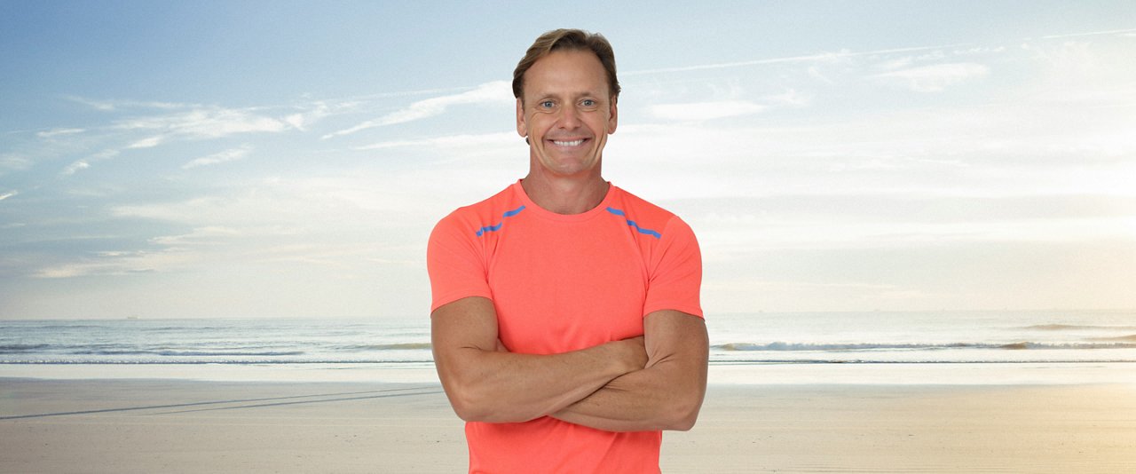 Guy Leech, AIA Ambassador, in an orange shirt stands on the beach, enjoying the sun and the ocean view.