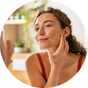 Woman touching her face and smiling, looking at her reflection in a sunlit bathroom