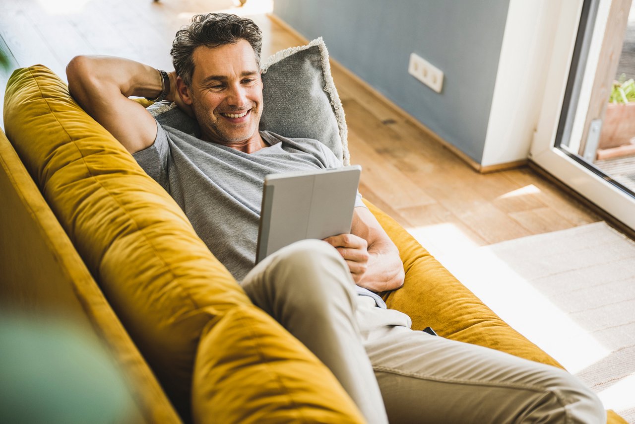Middle-aged man relaxing on his couch, smiling and looking at his tablet device.
