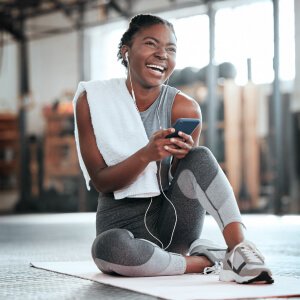 A woman sits on the floor with headphones and a phone, while a smiling man in gym attire holds a towel nearby.