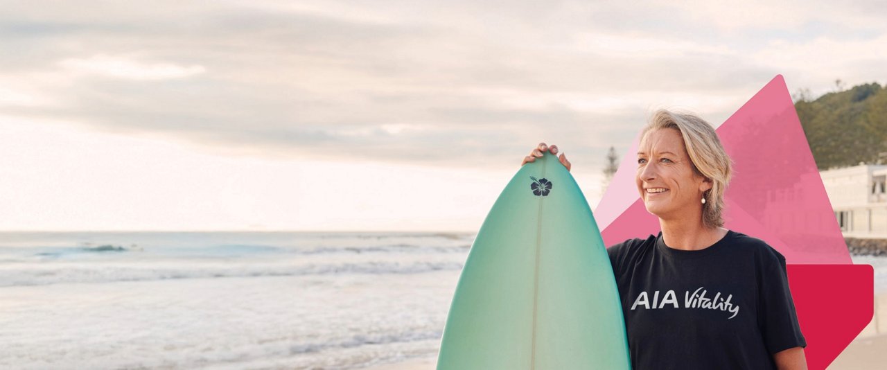 Layne Beachley, AIA Vitality Ambassador, stands on the beach holding a surfboard.