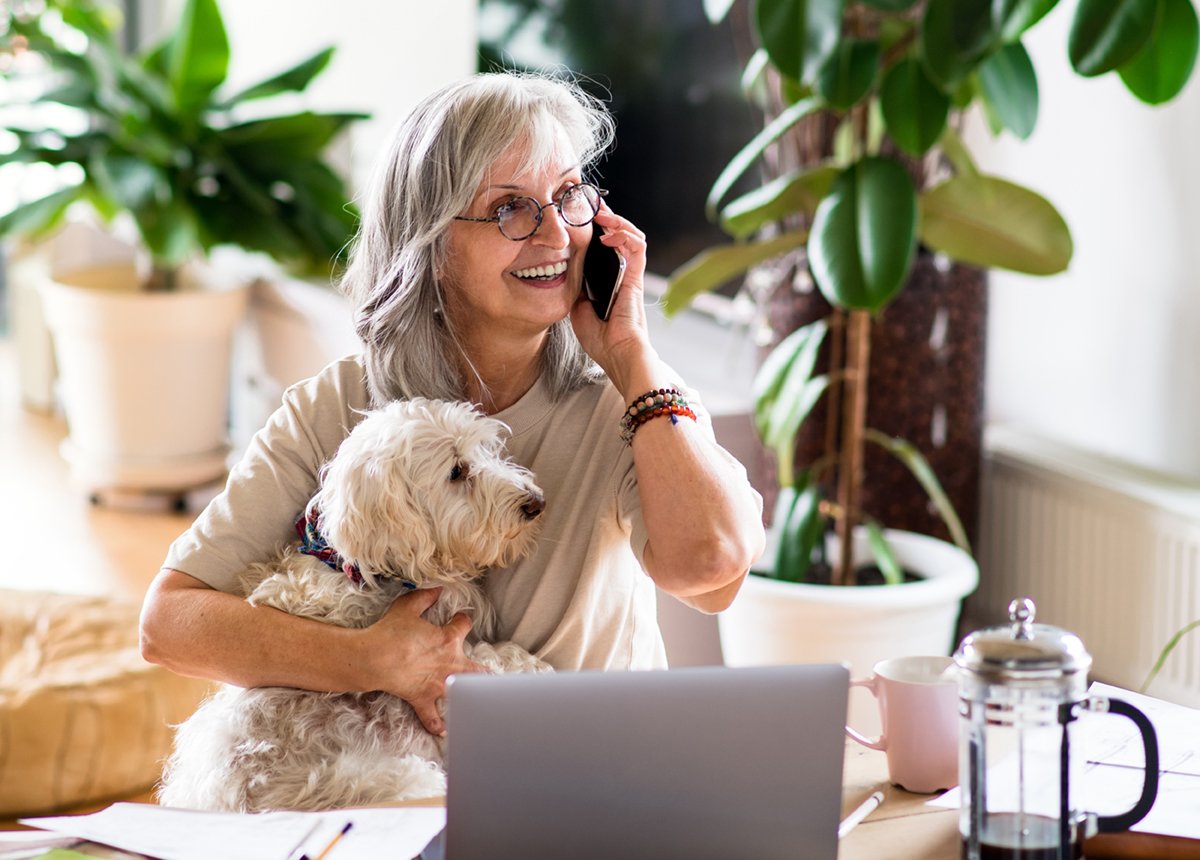 A woman wearing glasses is talking on the phone while her dog sits beside her