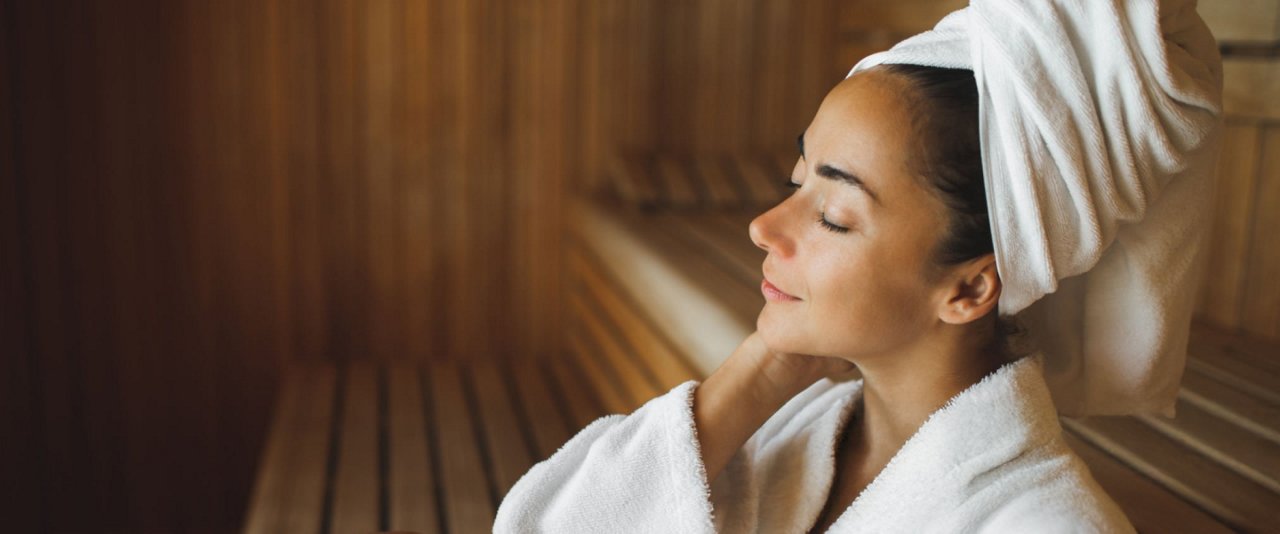 A woman wrapped in a towel relaxes peacefully in a warm sauna, surrounded by wooden benches and soft steam.