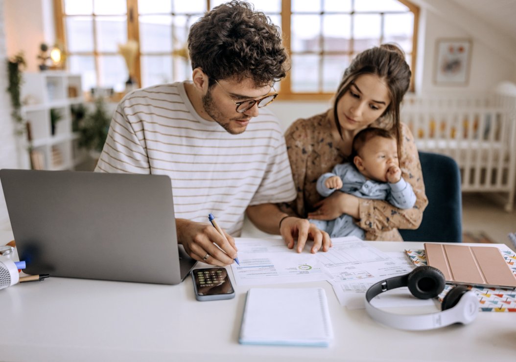 A man and woman collaborate on paperwork while gently cradling a baby in their arms.