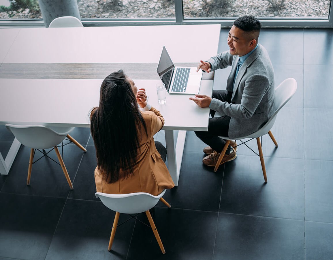 Two individuals seated at a table, each using a laptop, engaged in a collaborative work session.