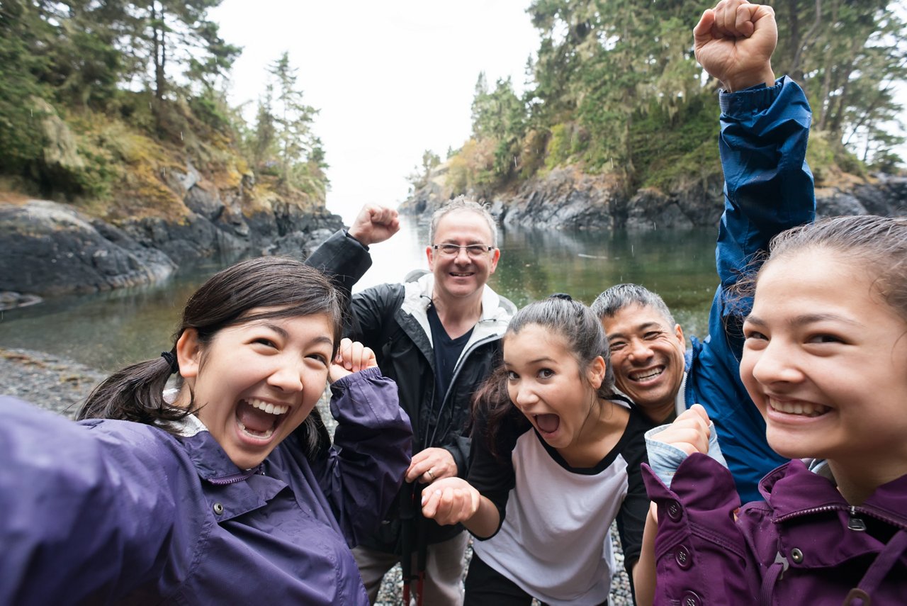 A real extended family of multi-ethnic and multi-generational backpackers in the wilderness pose of a selfie on a deserted beach surrounded by islands and forest with the ocean in the background.  Rainy day in a wilderness park.  Whiffen Spit, Sooke, British Columbia, Canada.