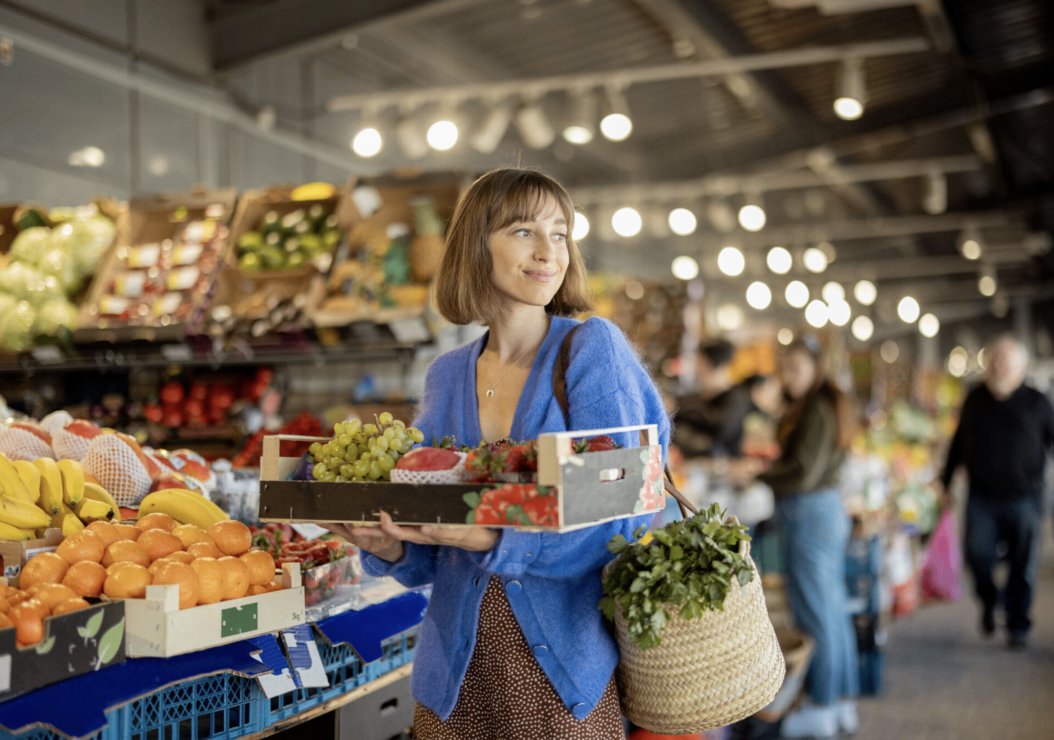 A woman stands in a grocery store, holding a basket filled with fresh fruits and vegetables.