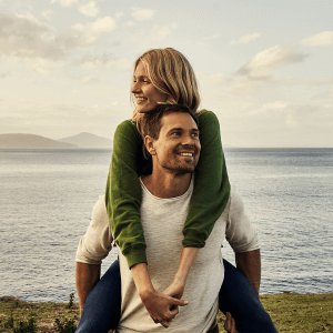 Man and woman smiling, the woman is riding piggyback on him, set against a scenic ocean backdrop