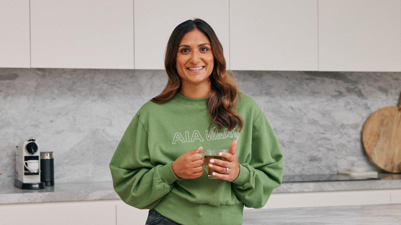 A woman in a green sweatshirt holds a cup of coffee, smiling as she enjoys her drink.